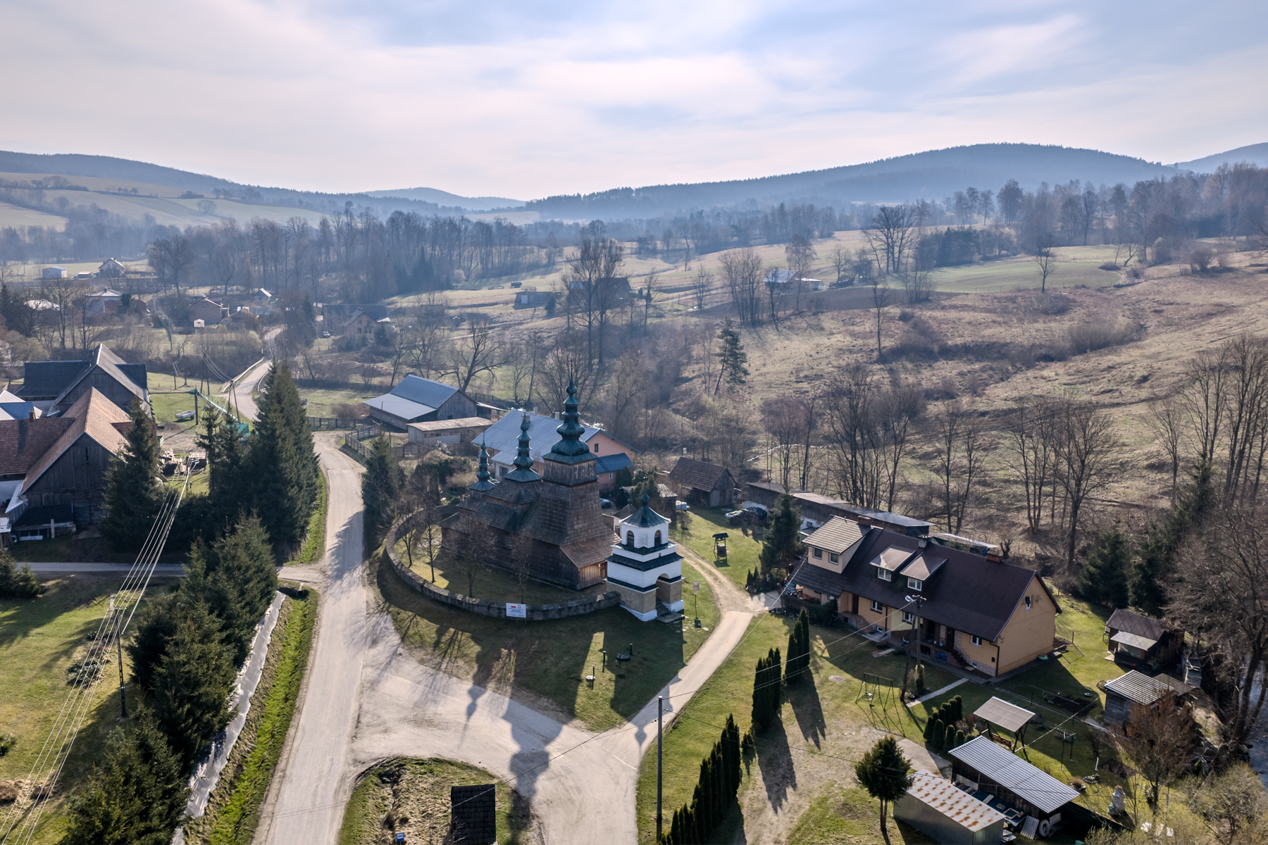 zdjęcie z drona przedstawiające budynek drewnianej cerkwi w Owczarach wraz z okolicznymi zabudowaniami na tle gór Beskidu Niskiego